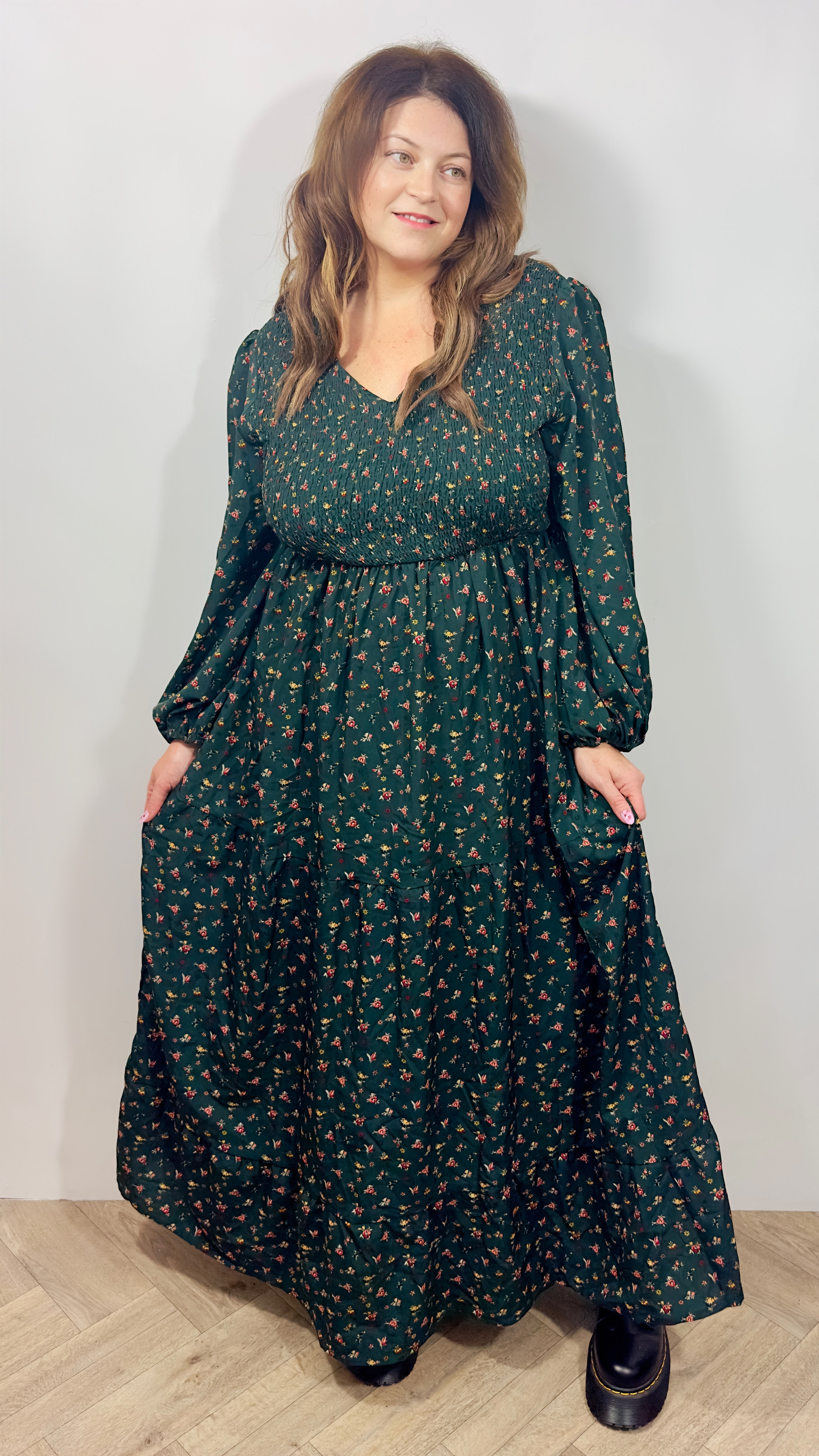 Woman wearing a long green floral dress against a white wall.