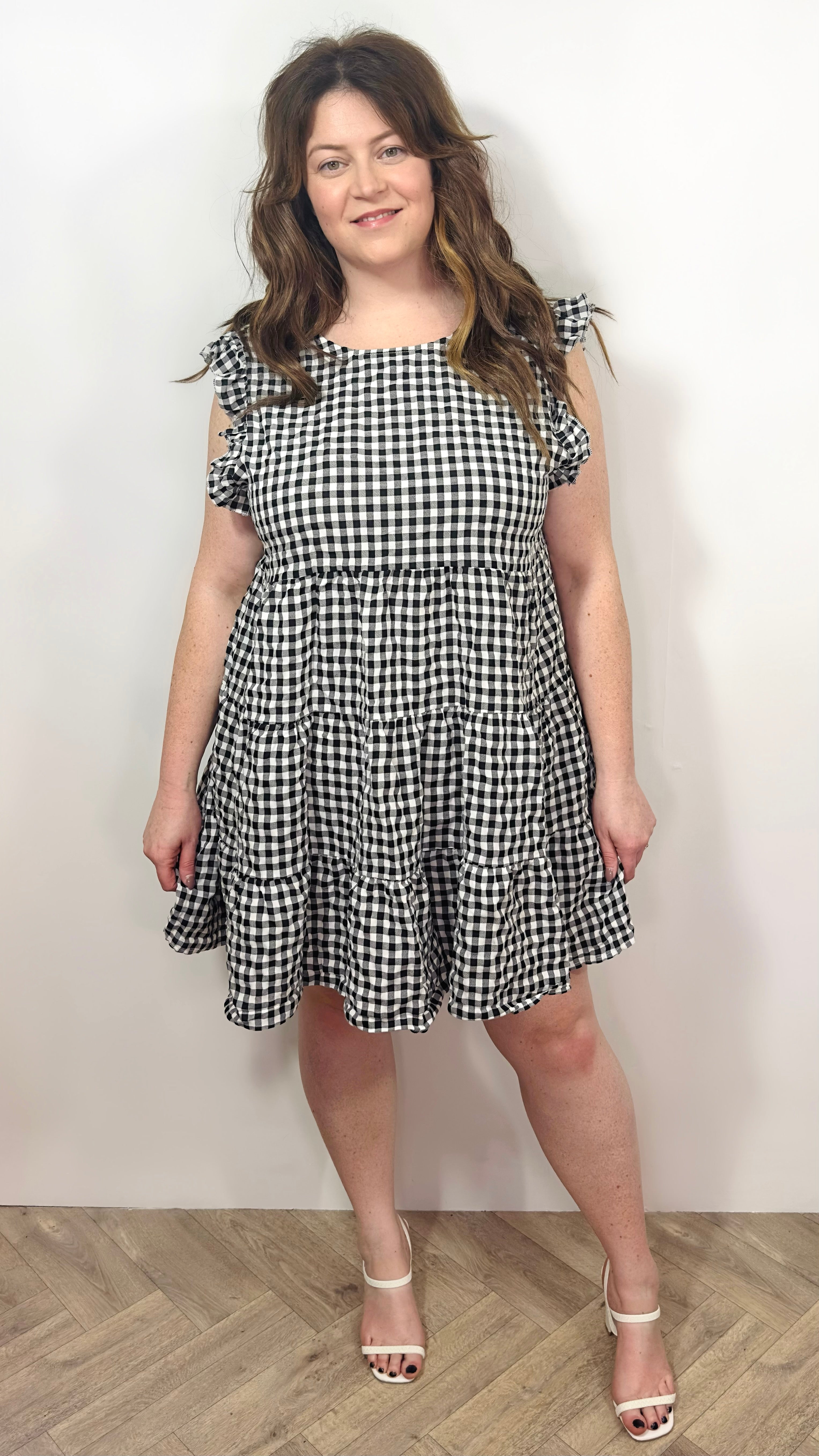 A model wearing a short-sleeved gingham smock dress with tiered detailing, standing against a plain background.