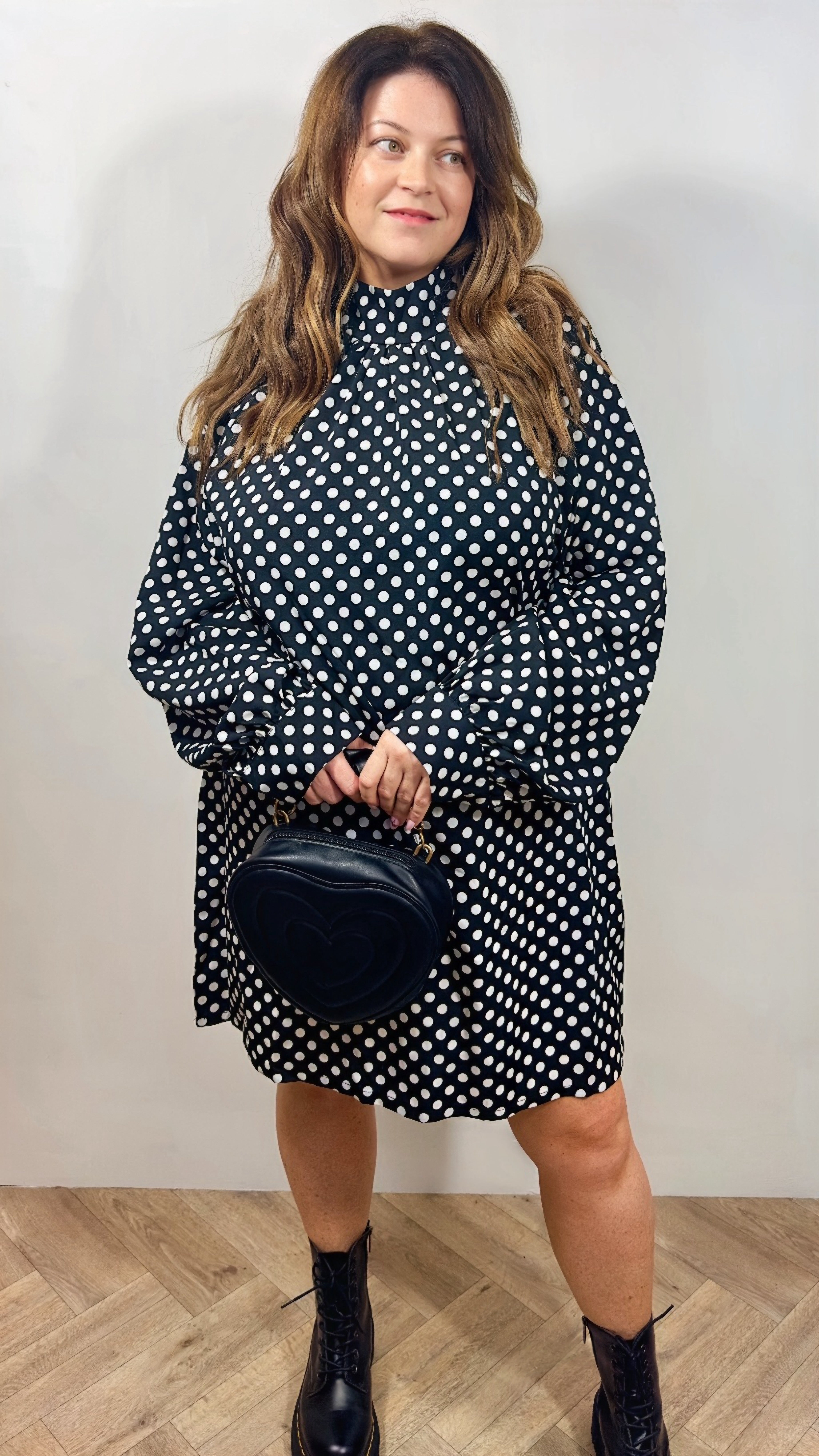 Woman wearing a black and white polka dot dress holding a black handbag against a plain background