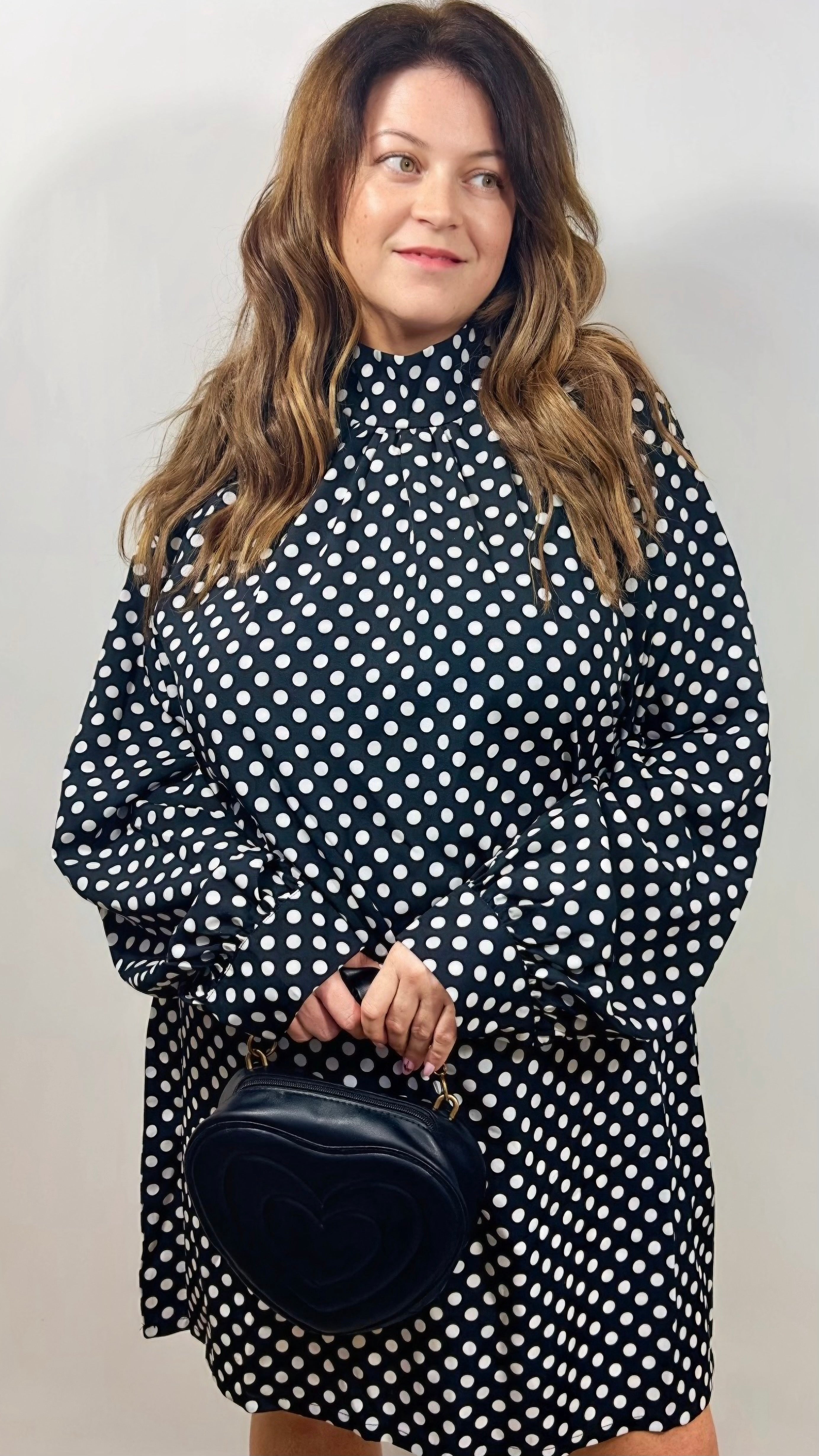 Woman wearing a black polka dot dress holding a black handbag against a plain background